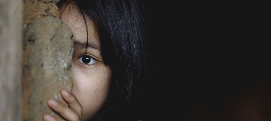 A child with dark hair peers from behind a stone wall, with one eye visible, set against a dark background.