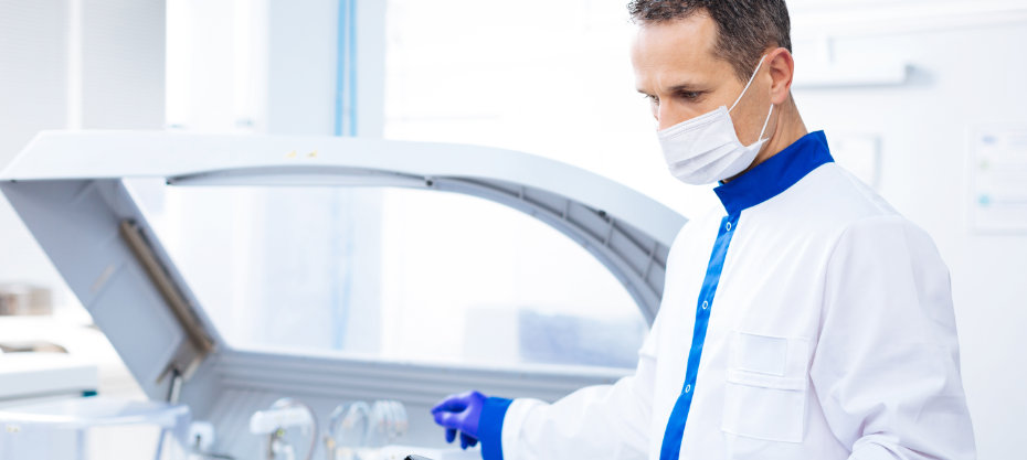 A male scientist in a lab coat and mask works with lab equipment, standing next to a large machine with an open lid.