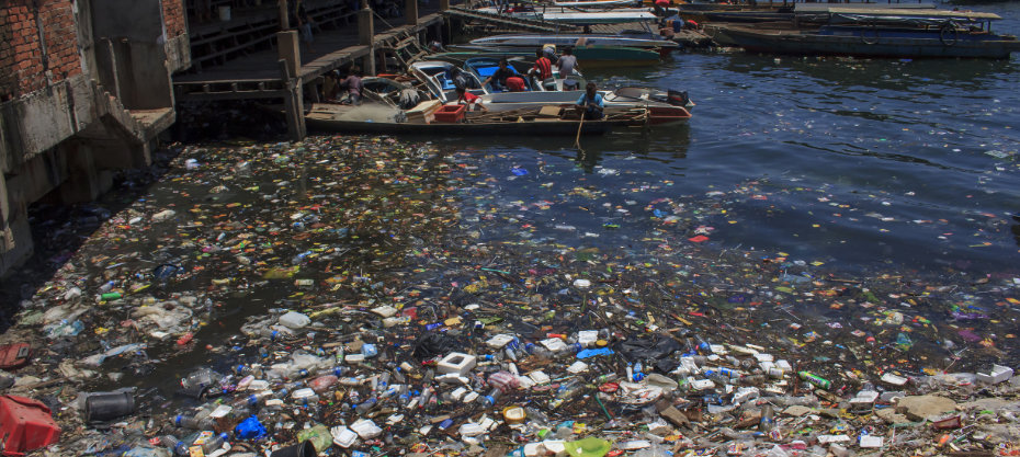 A heavily polluted waterbody near a dock, littered with plastic and trash, with boats moored nearby.